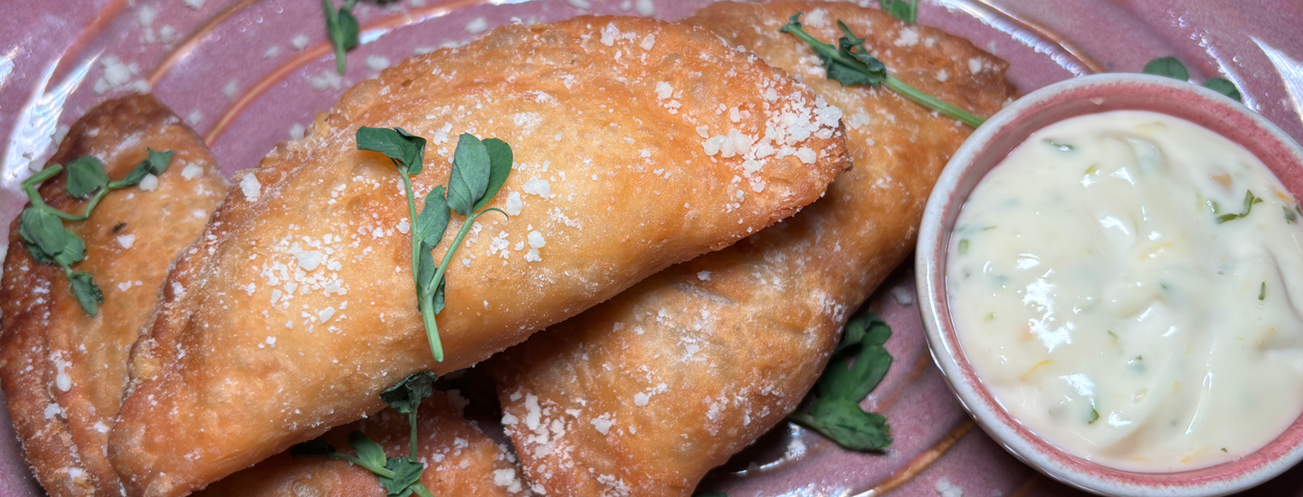 Three golden brown empanadas with garnish and dipping sauces on a soft pink colored plate.